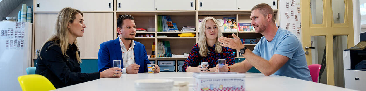 Mariella van Schie, Jerry Straten, Carmen Massaar en René Rusman zitten samen aan een tafel in een klaslokaal. René praat en gebruikt daarbij zijn handen, de andere drie kijken hem aan.