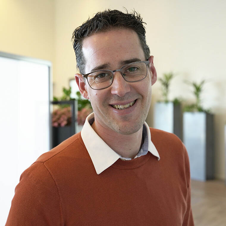 Pieter van der Bent is een witte man met donker haar en een bril. Hij draagt een oranje trui, leunt iets naar de camera en lacht.