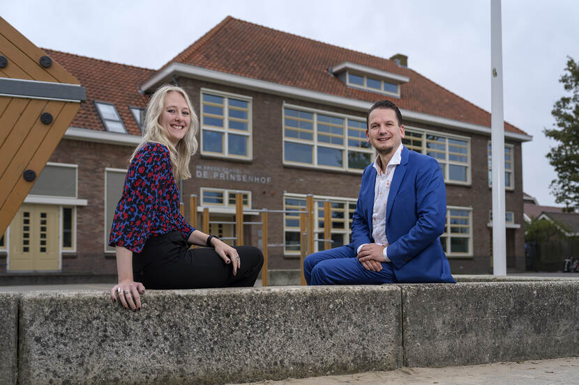 Carmen Massaar is een blonde vrouw met een blauw met rode bloes. Jerry Straten is een witte man met donkerblond haar en een blauw pak. Samen zitten ze op een muurtje, achter hen is basisschool De Prinsenhof in Noordwijkerhout te zien.