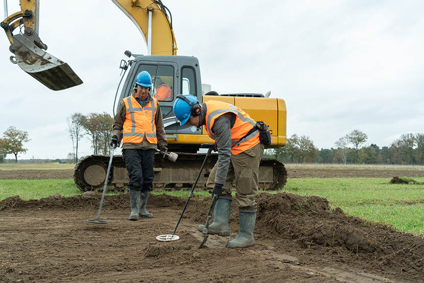 Twee archeologen met oranje hesjes en een graafmachine op de achtergrond, zoekende naar materiaal in de grond