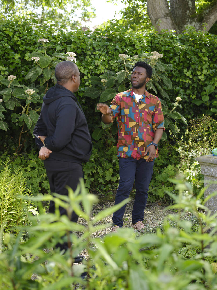 Marcel Pinas staat links, in het zwart gekleed, we zien hem van op zijn rug. Hij is in gesprek met Isan Corinde, die een kleurrijke bloes aan heeft. Ze staan in een park of soortgelijke groene omgeving.