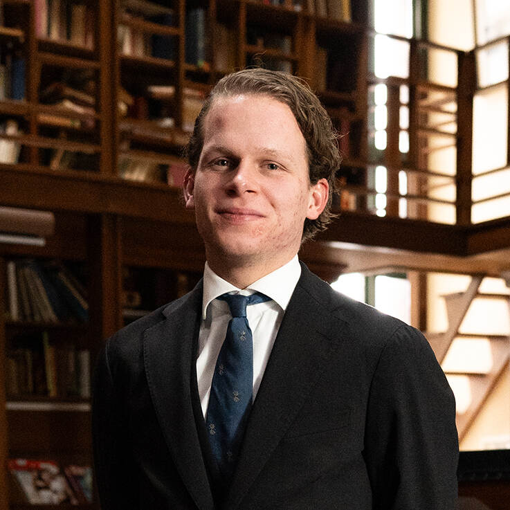 Portret Reinier Nijman. Hij staat in oude bibliotheek en kijkt in de camera. Hij is gekleed in nette kleding